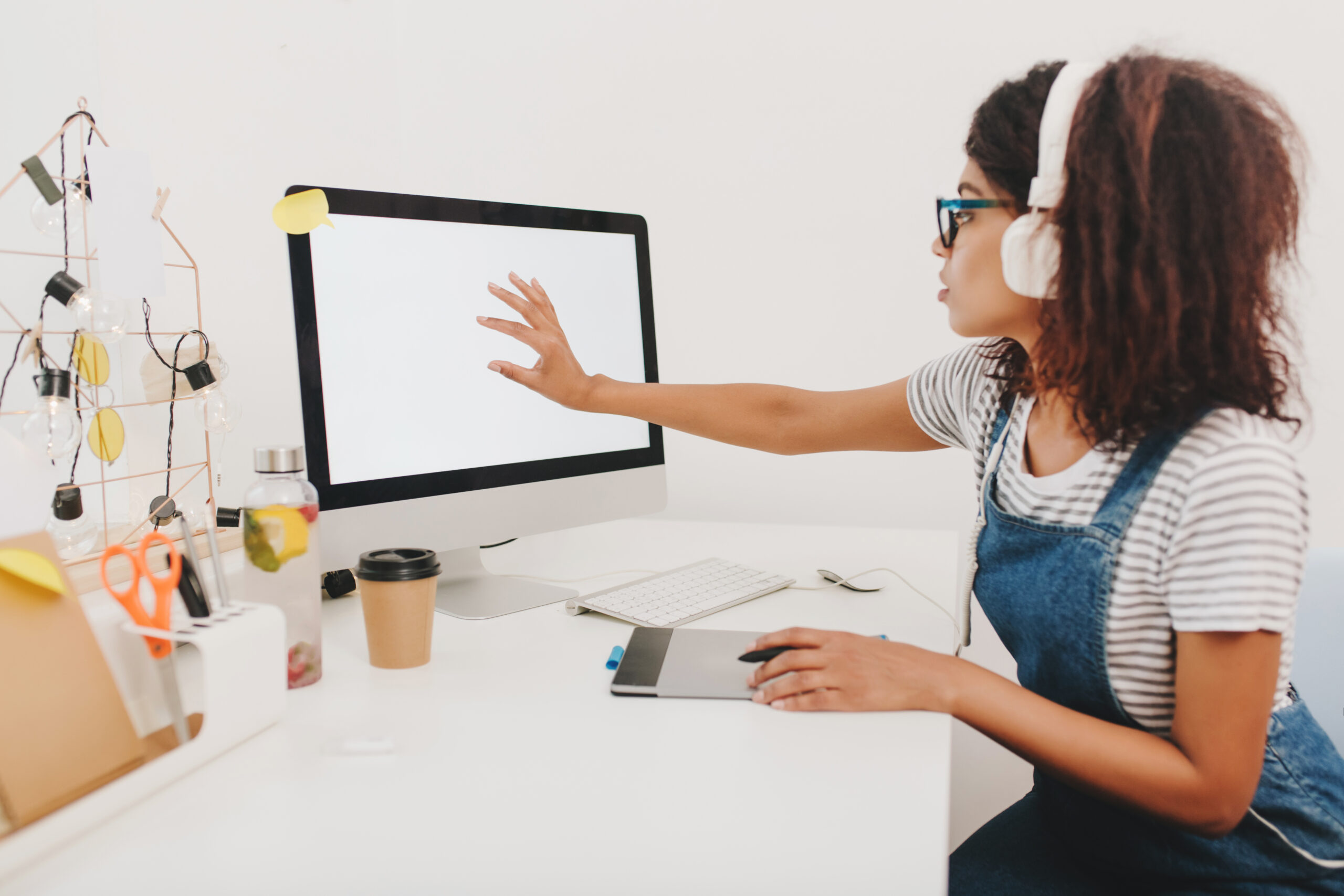 Black girl in denim sitting at the table with stationery and touching computer screen. Indoor portrait of pretty young woman in headphones with tablet spending time in office..
